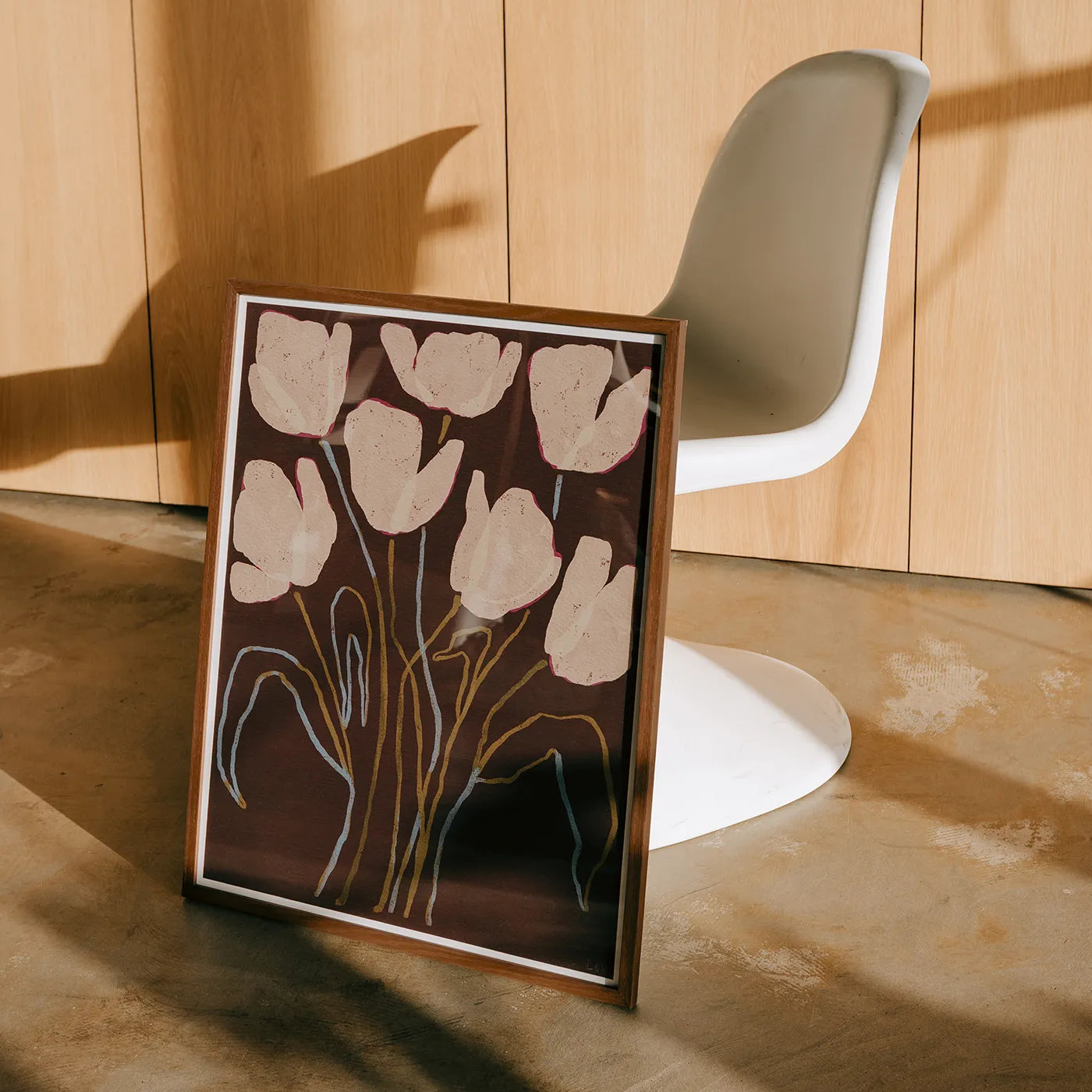 A framed floral art print leaning against a wall in a modern interior, featuring cream tulip-like flowers on a dark brown background with gold and white line details. A white molded chair and light wood paneled wall are visible behind it.