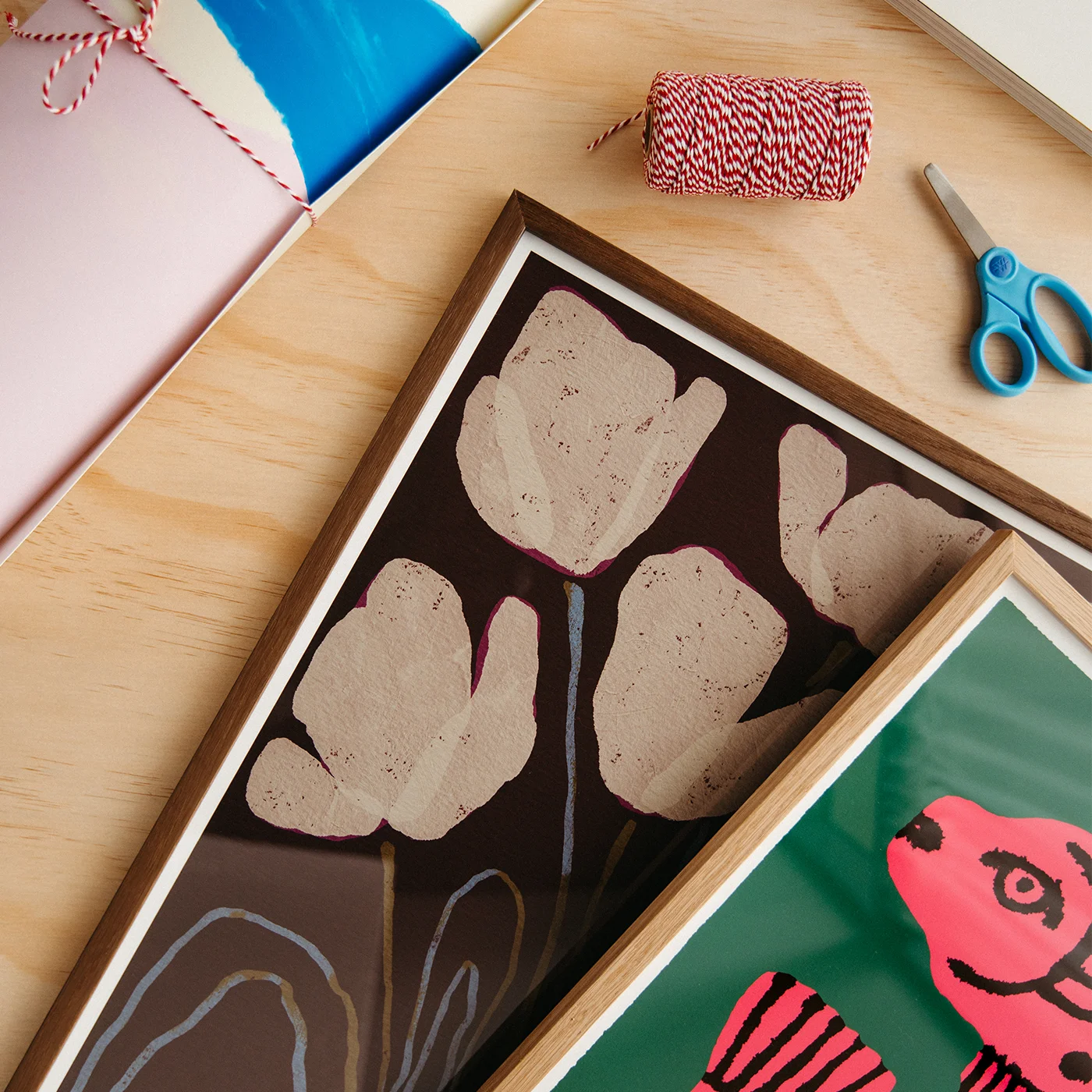 A styled flat-lay of framed art prints on a light wood surface, alongside a pink gift box, blue-handled scissors, and a spool of red-and-white striped twine. The main featured print shows large beige tulip-like floral shapes on a dark brown background, with a second partial print below showing a bright pink fish on green.
