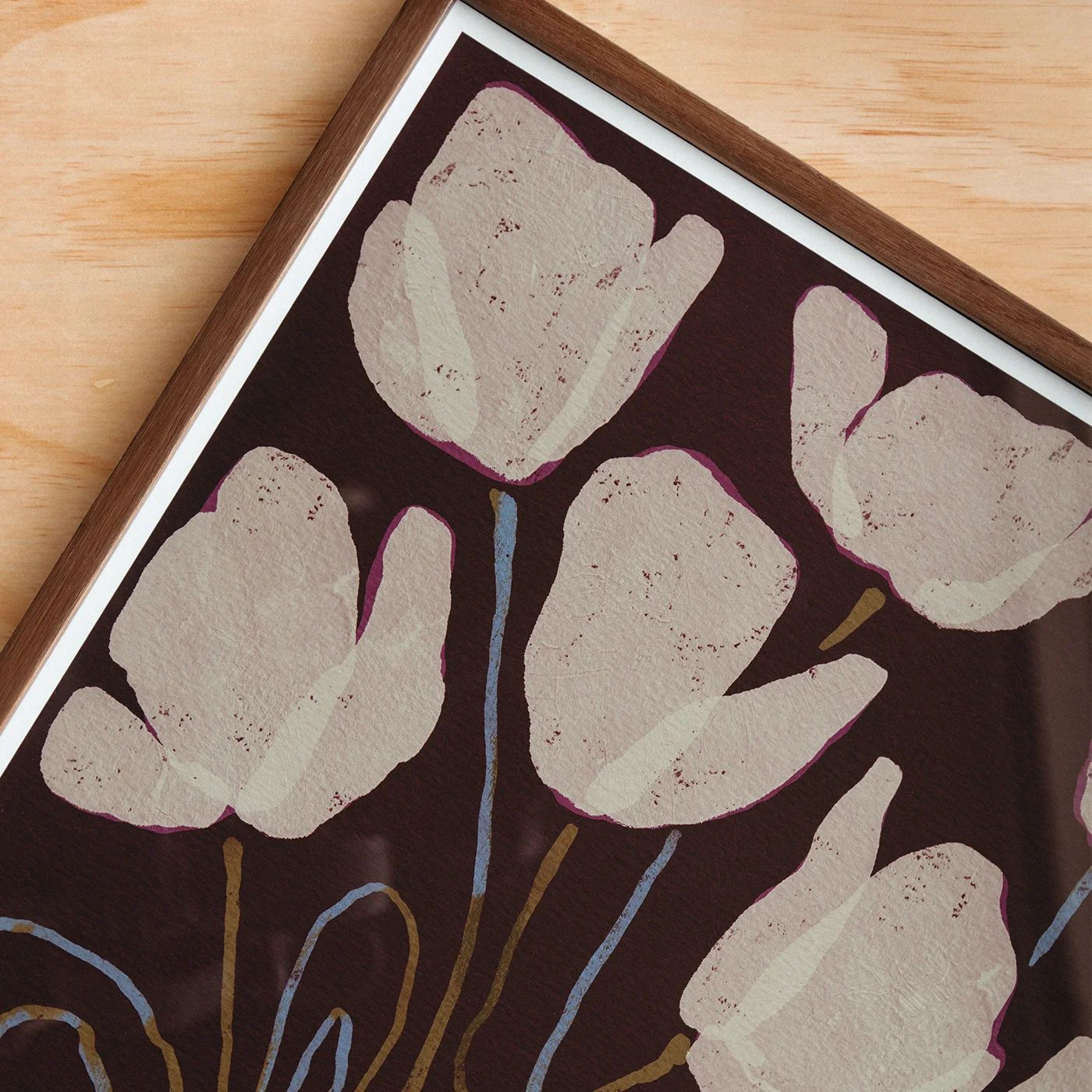 Framed floral wall art featuring large pale blush tulip-like shapes on a dark brown background, with blue and tan stems, shown in a wood frame on a light wood surface.