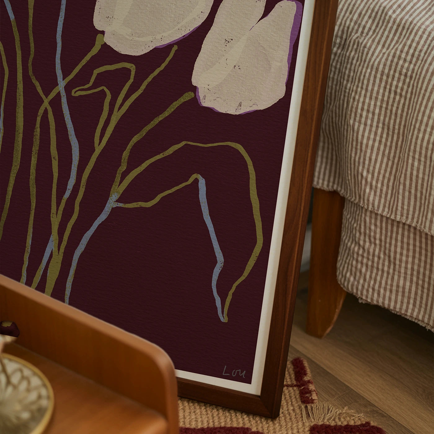 A framed abstract floral art print with cream tulip-like shapes and olive and light blue stems on a deep purple background, leaning against a bed in a bedroom setting.