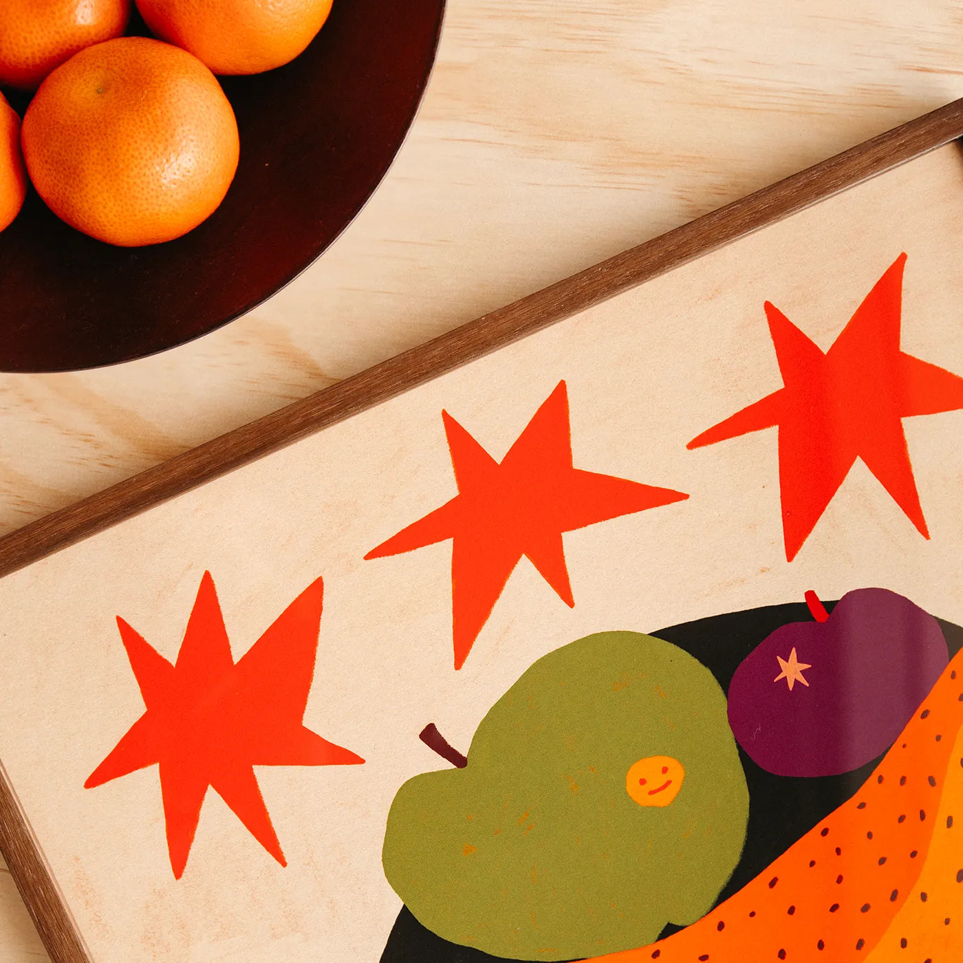 A close-up of a framed art print with a colorful fruit and star artwork, styled on a light wood surface with oranges in a dark bowl.