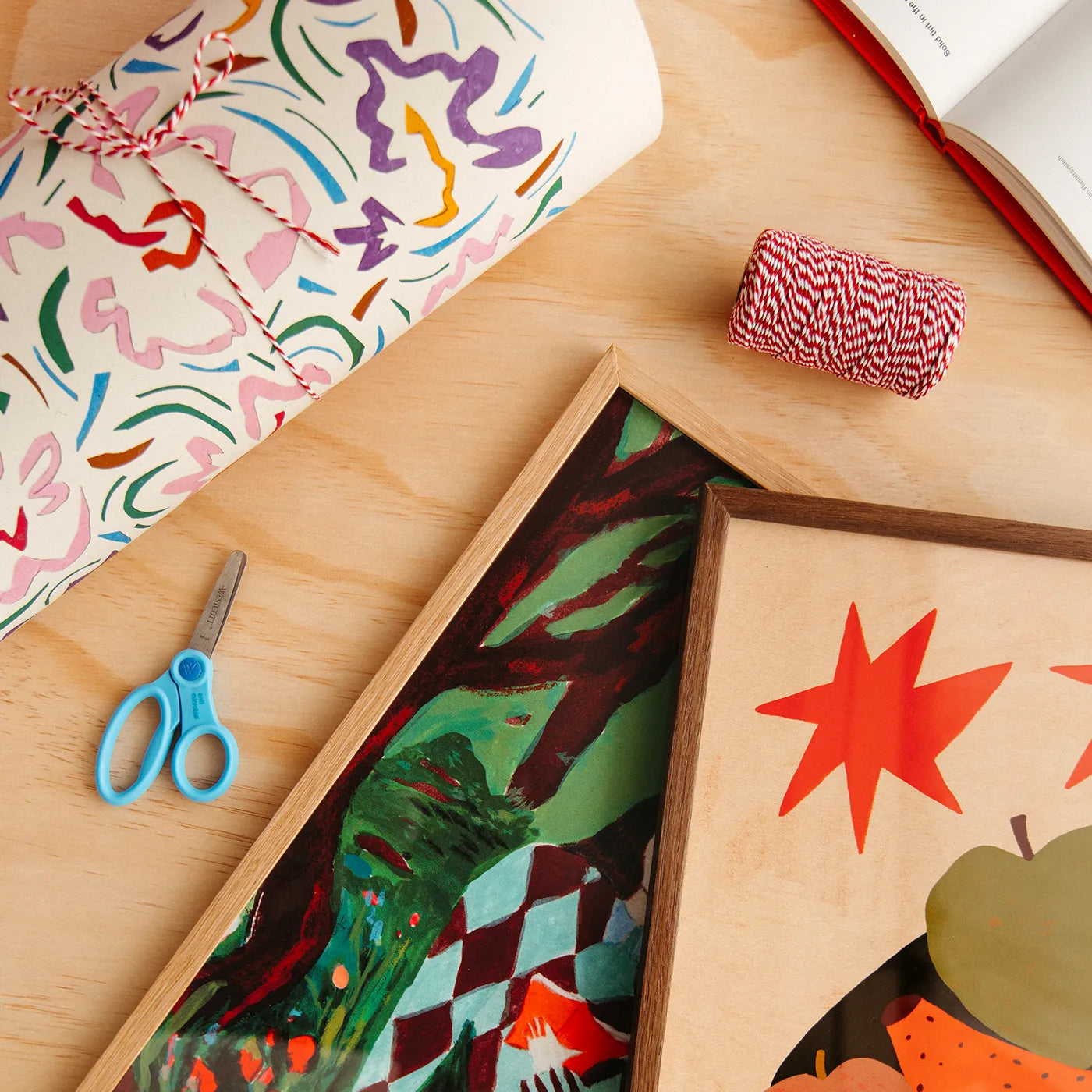 A styled flat-lay of wrapping paper and gift-wrapping supplies on a light wood surface, including a rolled sheet with colorful abstract print, a spool of red-and-white patterned twine, blue-handled scissors, and framed artwork.