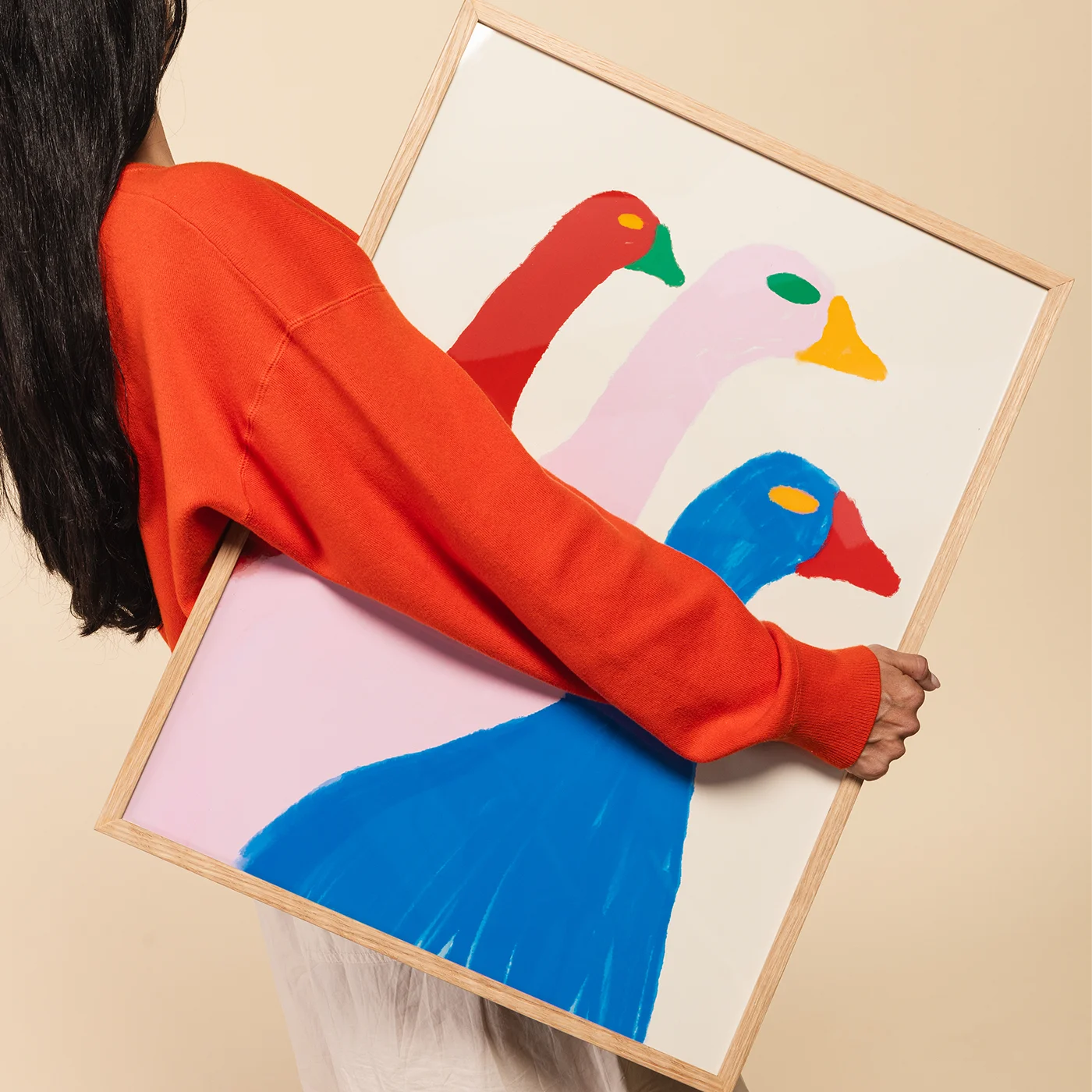 A person in a bright orange sweatshirt holds a light wood framed art print featuring three stylized ducks in red, pink, blue, and yellow on a cream background.