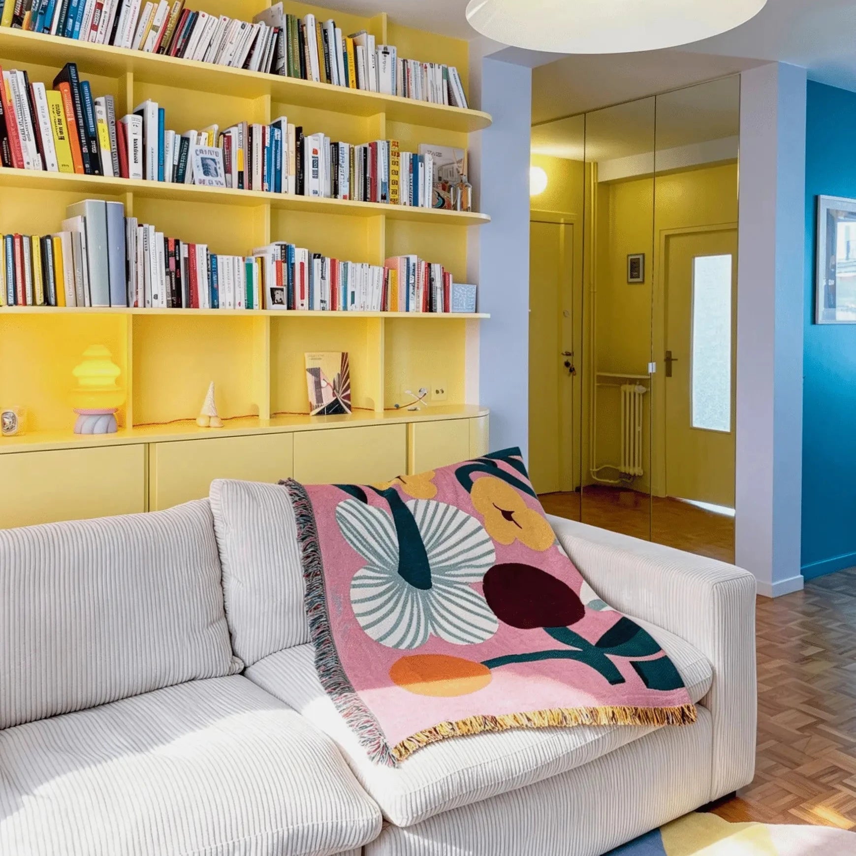 A cozy living room scene featuring a white ribbed sofa with a pink, yellow, and blue floral patterned throw blanket draped over it, set against a bright yellow bookshelf and yellow walls.