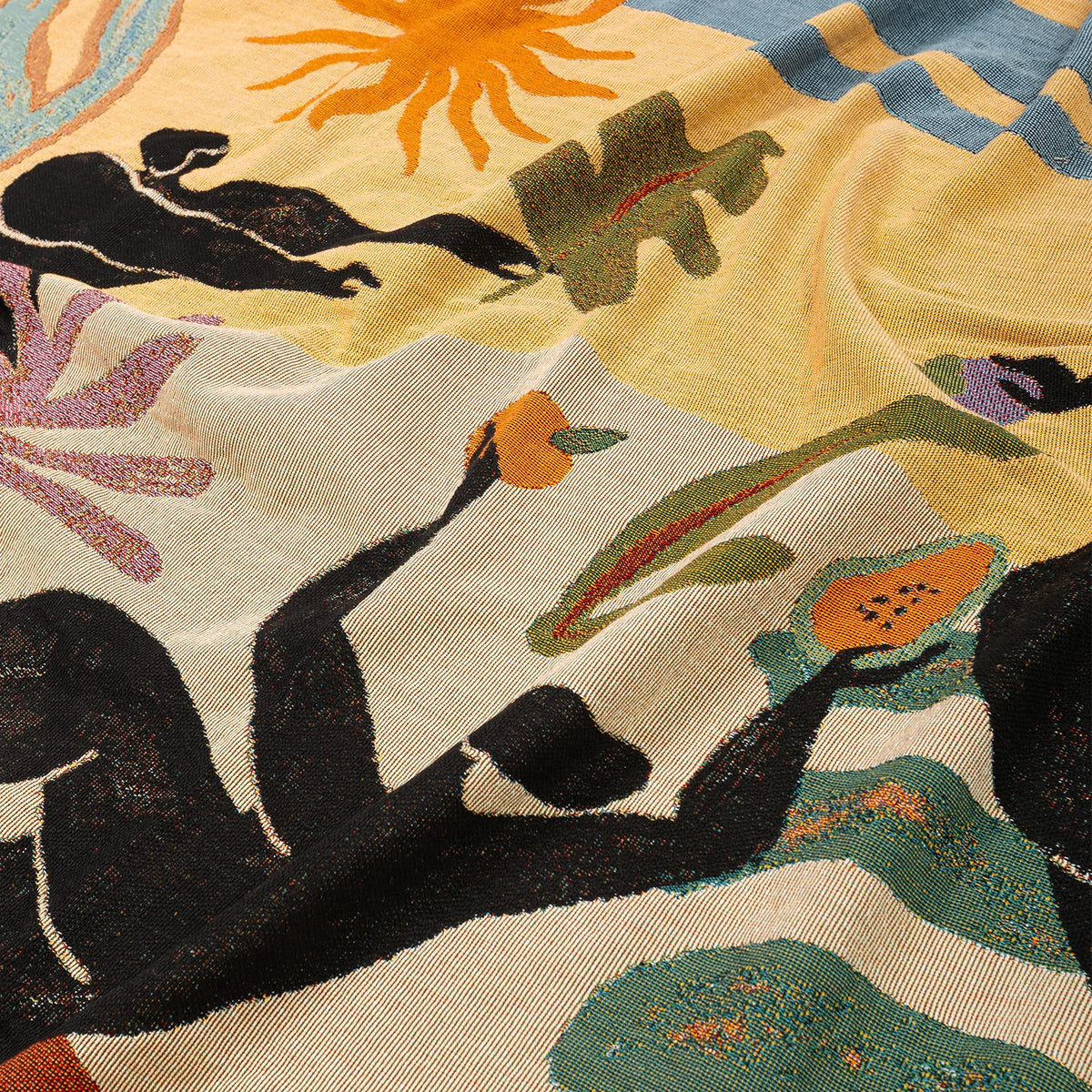 A close-up of a woven blanket featuring a colorful abstract and botanical pattern with black silhouetted figures, green leaves, orange sun and fruit, and purple floral elements on a cream and pale yellow background.