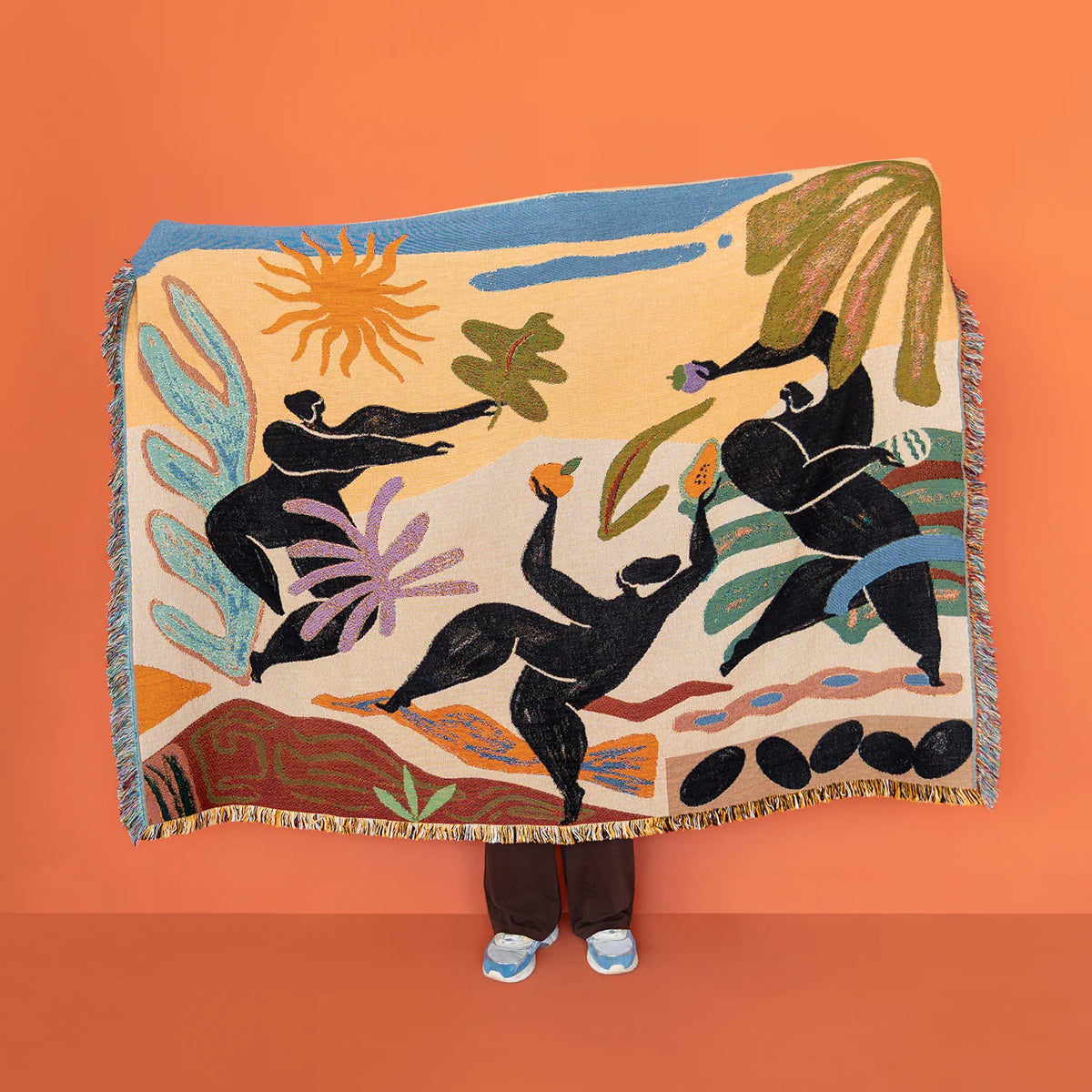 A large woven throw blanket with fringe is held up against a solid orange background. The blanket features a colorful, abstract folk-art style design with black human figures, a yellow sun, and green and blue botanical elements on a cream base.