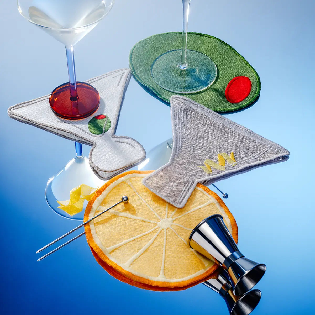 Set of linen drink coasters shaped like a martini glass, orange slice, and cocktail shaker on a blue background.