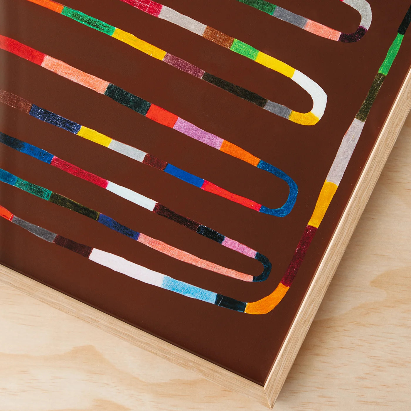 A close-up of a framed art print featuring a brown background with wavy, snake-like multicolor lines, resting on a light wood surface.