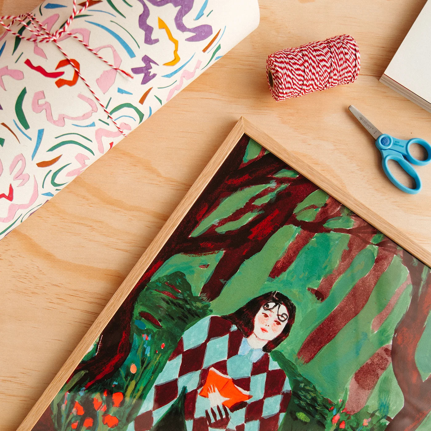 A framed art print depicting a woman in a red and white checkered coat holding an orange book in a forest, styled on a light wood table with a wrapped gift, bakers twine, and blue-handled scissors.