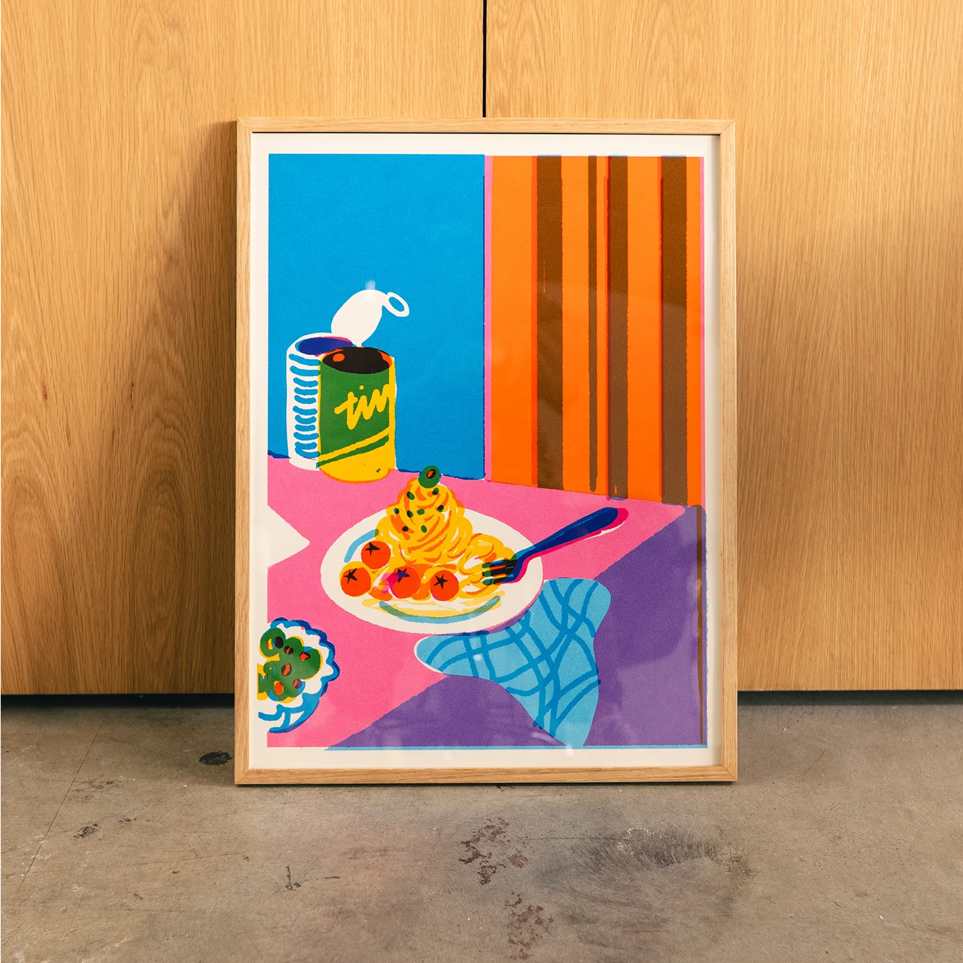 A framed pop art print depicting a colorful still life of spaghetti, tomatoes, broccoli, and a can of soup, leaning against a light wood-paneled wall on a concrete floor.