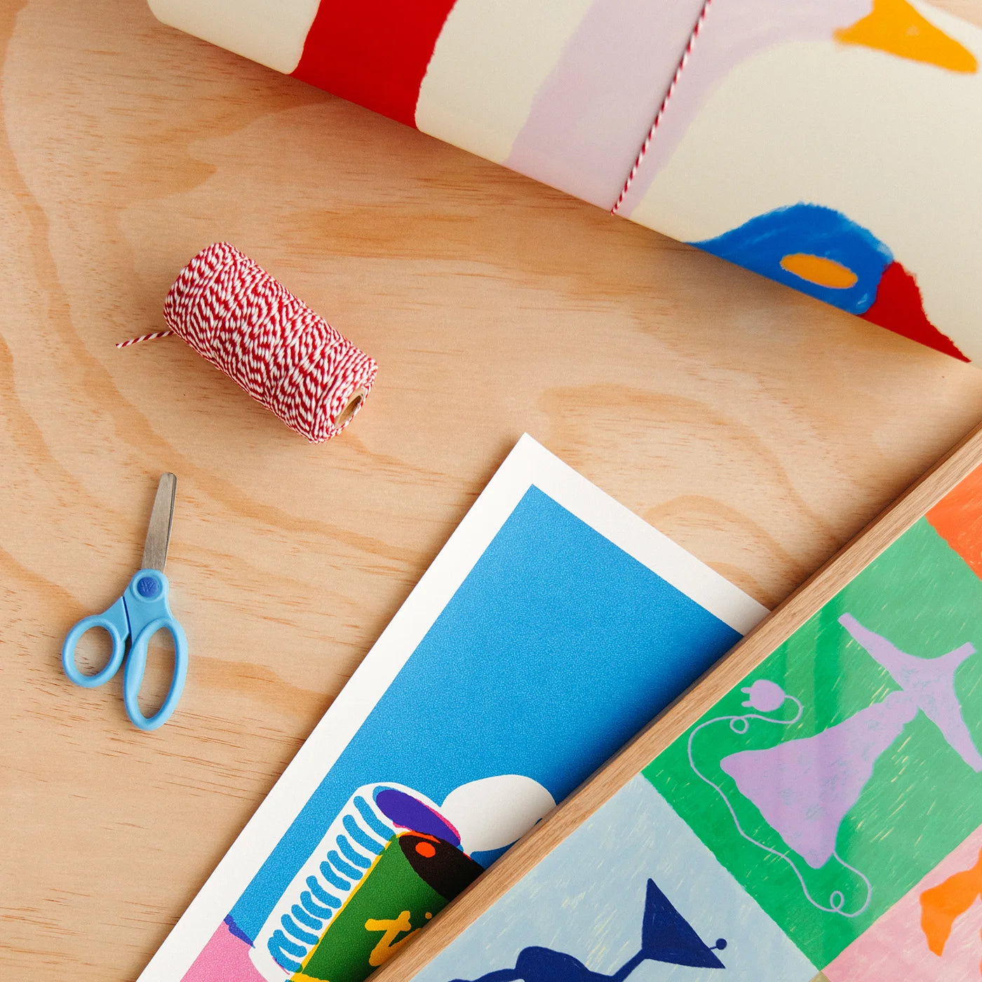A styled flat-lay of a colorful patterned fabric or paper roll, a spool of red-and-white bakers twine, blue-handled scissors, and art prints on a light wood surface.