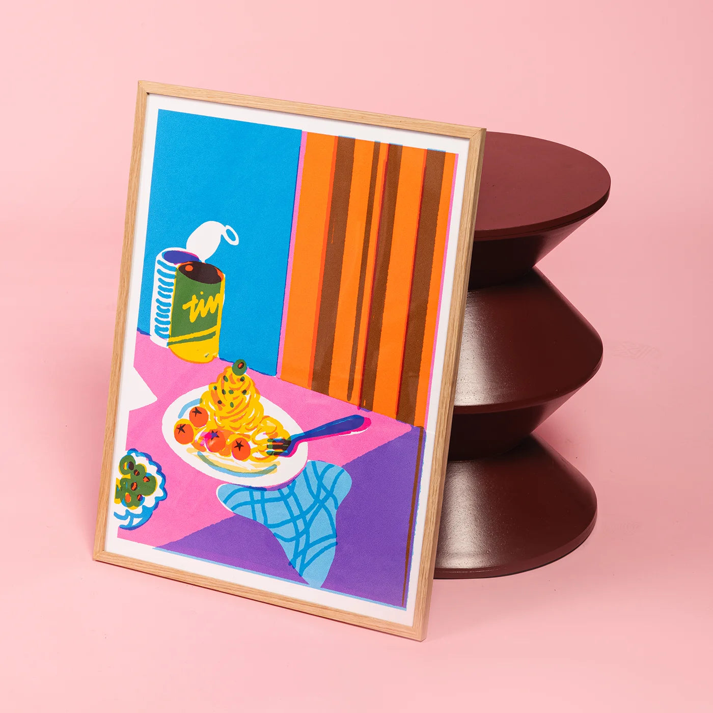 A framed art print featuring a colorful still life of spaghetti with tomatoes, a blue napkin, and a yellow can, leaning against a dark brown sculptural stand on a pink background. The wall art is framed in oak timber.