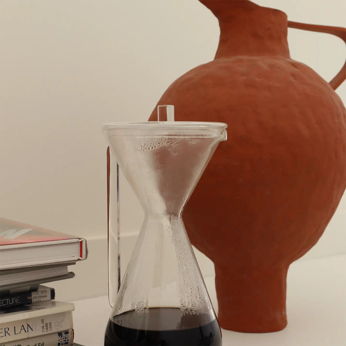 Hourglass clear carafe next to a terracotta pot on a surface.