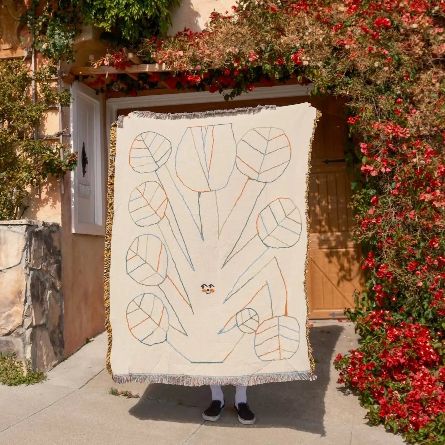 Person holding a decorative blanket with leaf patterns in front of a building with flowers.