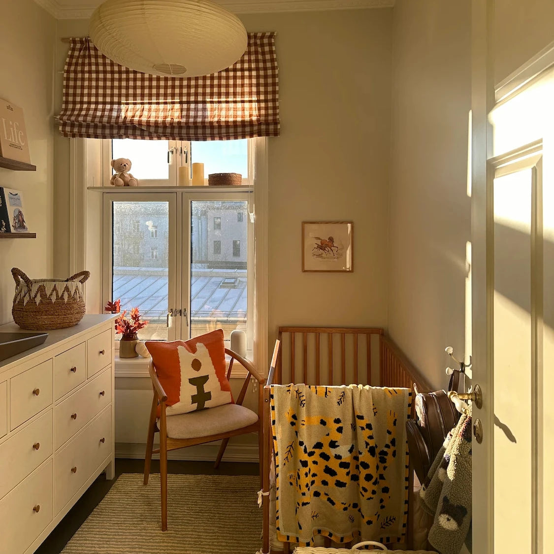 Nursery with checkered curtain, crib, and decorative items. A blanket with a tiger animal design hangs over the cot.