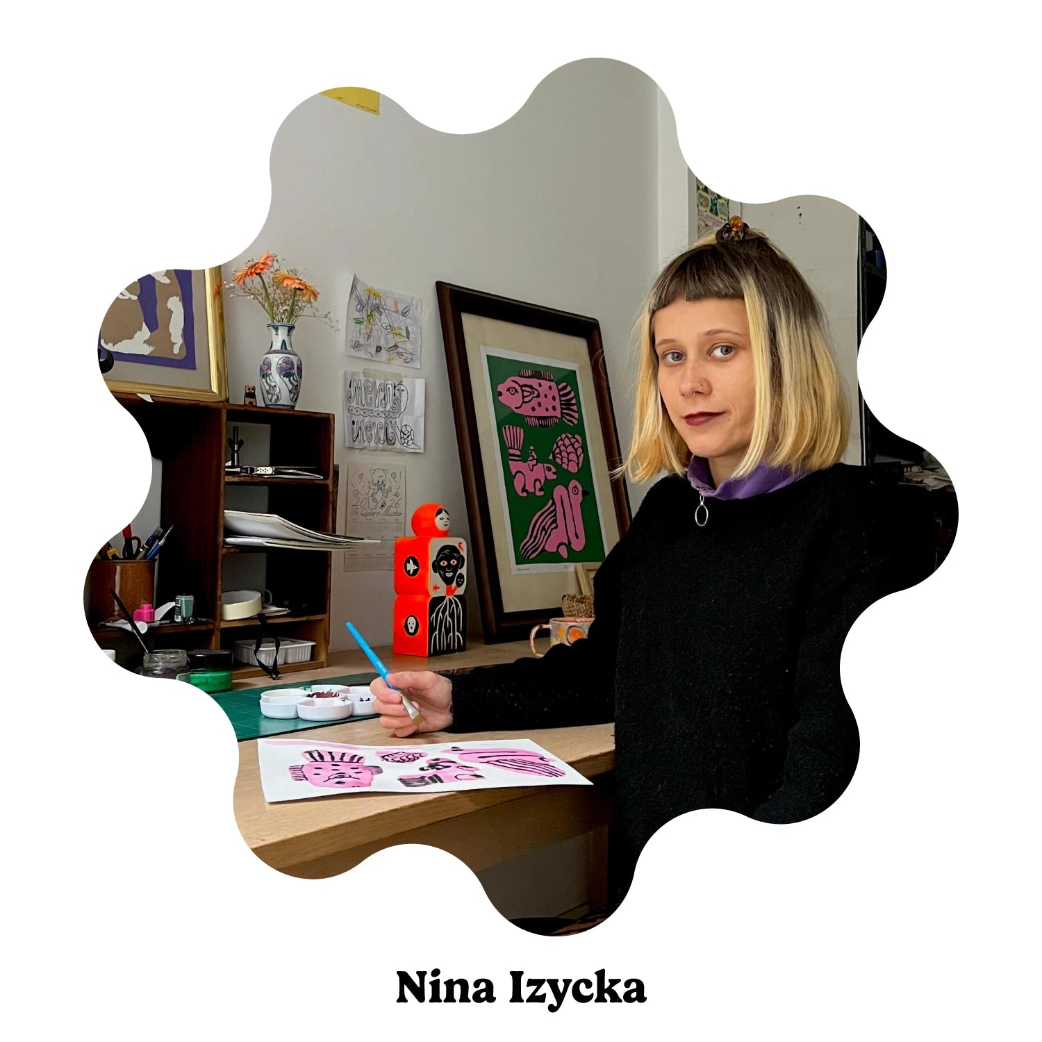 Portrait of Spanish artist Nina Izycka seated at a desk, holding a blue pen and working on a pink artwork. The scene includes framed art, shelves, and a vase with flowers.