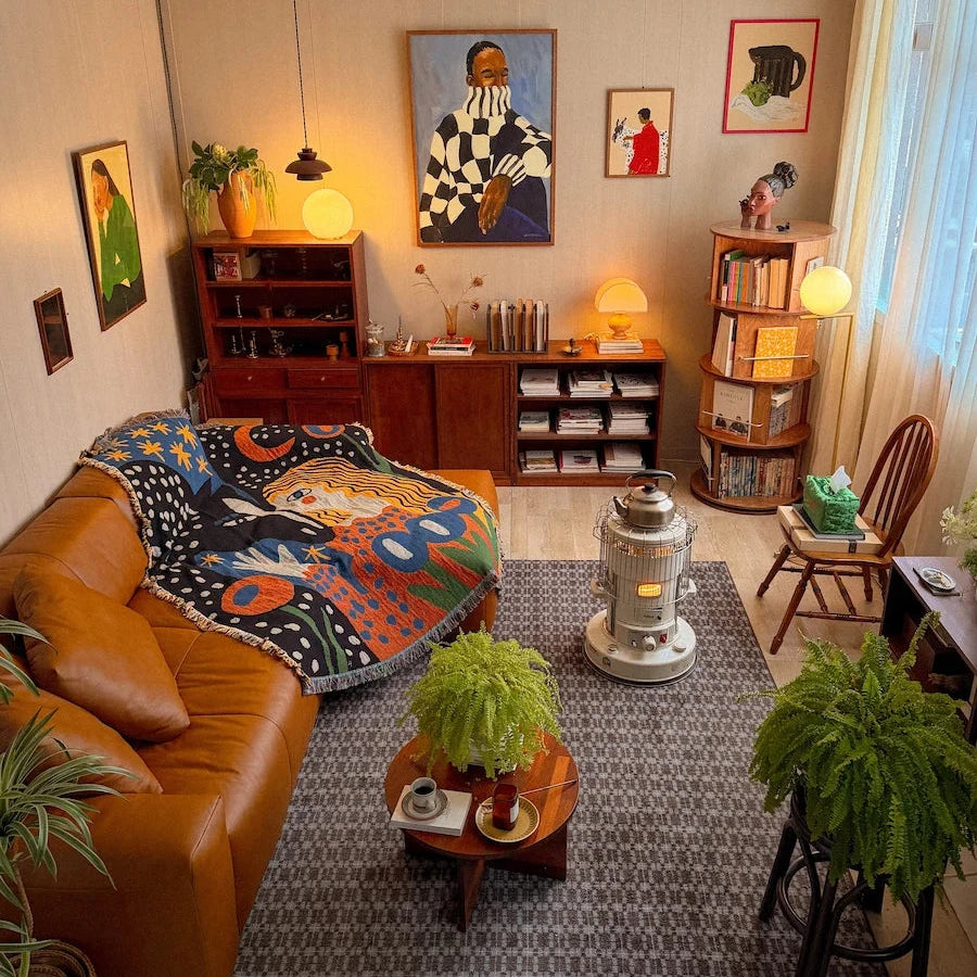 A cozy, vintage-style living room interior featuring a brown leather sofa with a colorful patterned blanket, a gray patterned rug, a small wooden coffee table, potted ferns, bookshelves, and framed art on the beige walls.