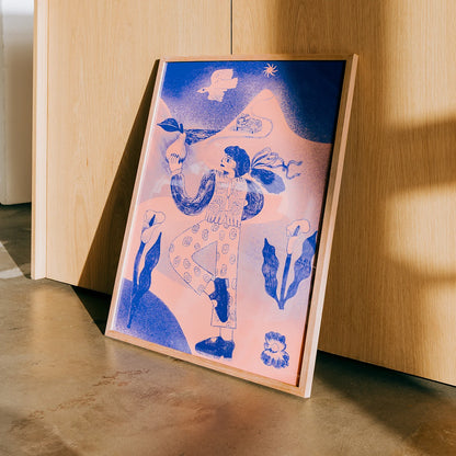 A framed art print leaning against a light wood cabinet on a concrete floor. The print features a stylized blue and pink illustration of a woman in a patterned dress against a mountain background.