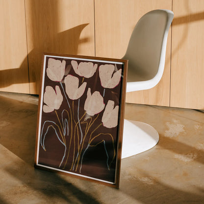 A framed floral art print leaning against a wall in a modern interior, featuring cream tulip-like flowers on a dark brown background with gold and white line details. A white molded chair and light wood paneled wall are visible behind it.
