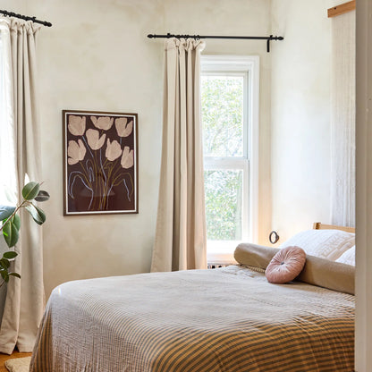 Bedroom wall art featuring a framed floral art print in a styled bedroom interior featuring beige curtains hanging from a black rod, a bed with beige and brown striped bedding, a pink round pillow.