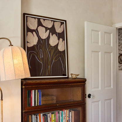 Bedroom wall art featuring cream tulips on a dark brown background, framed in dark walnut wooden frame. The art print is displayed on a wooden bookcase in a bedroom next to a lit lamp and an open white door.