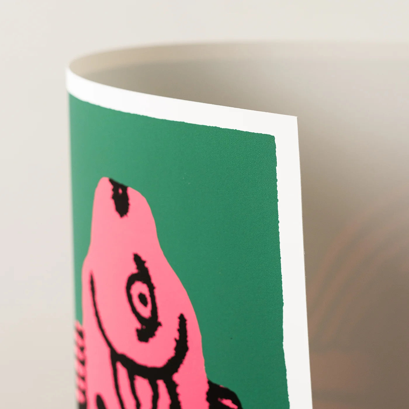 A close-up of an art print showing a printed graphic with a pink abstract shape outlined in black on a green background, photographed against a soft beige studio backdrop.