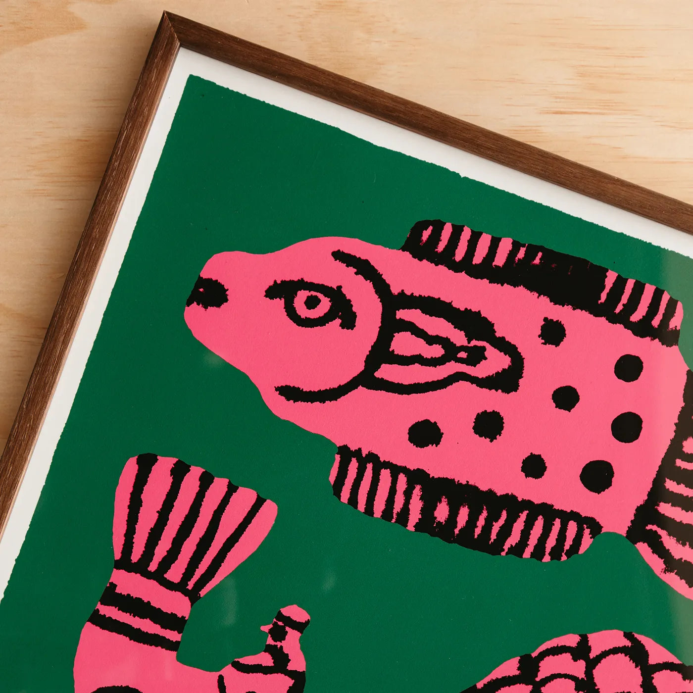 A framed art print featuring a stylized pink fish with black spots and stripes on a dark green background, shown at an angle on a light wood surface.
