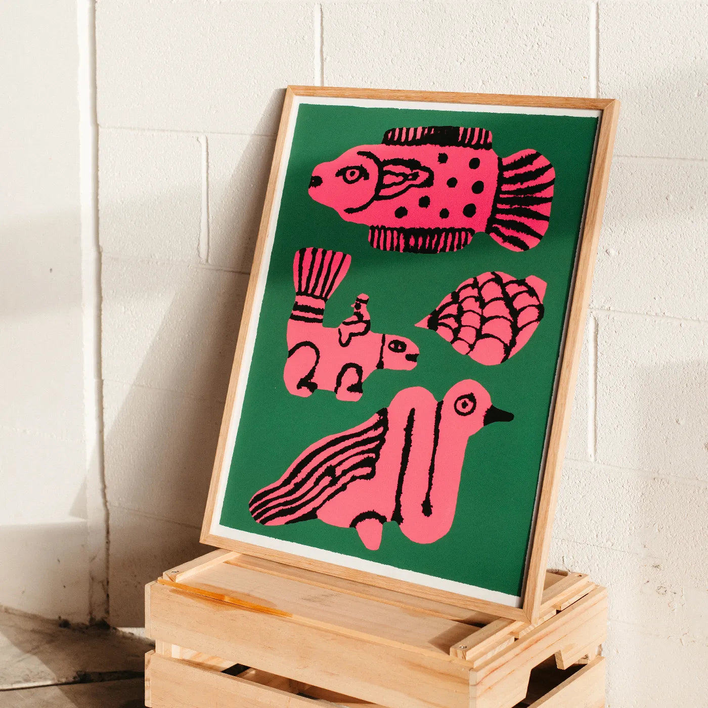 A framed art print featuring stylized pink fish, a shell, and a bird on a dark green background, leaning on a wooden crate against a white painted brick wall.