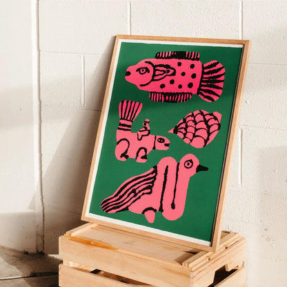 A framed art print featuring stylized pink fish, a shell, and a bird on a dark green background, leaning on a wooden crate against a white painted brick wall.