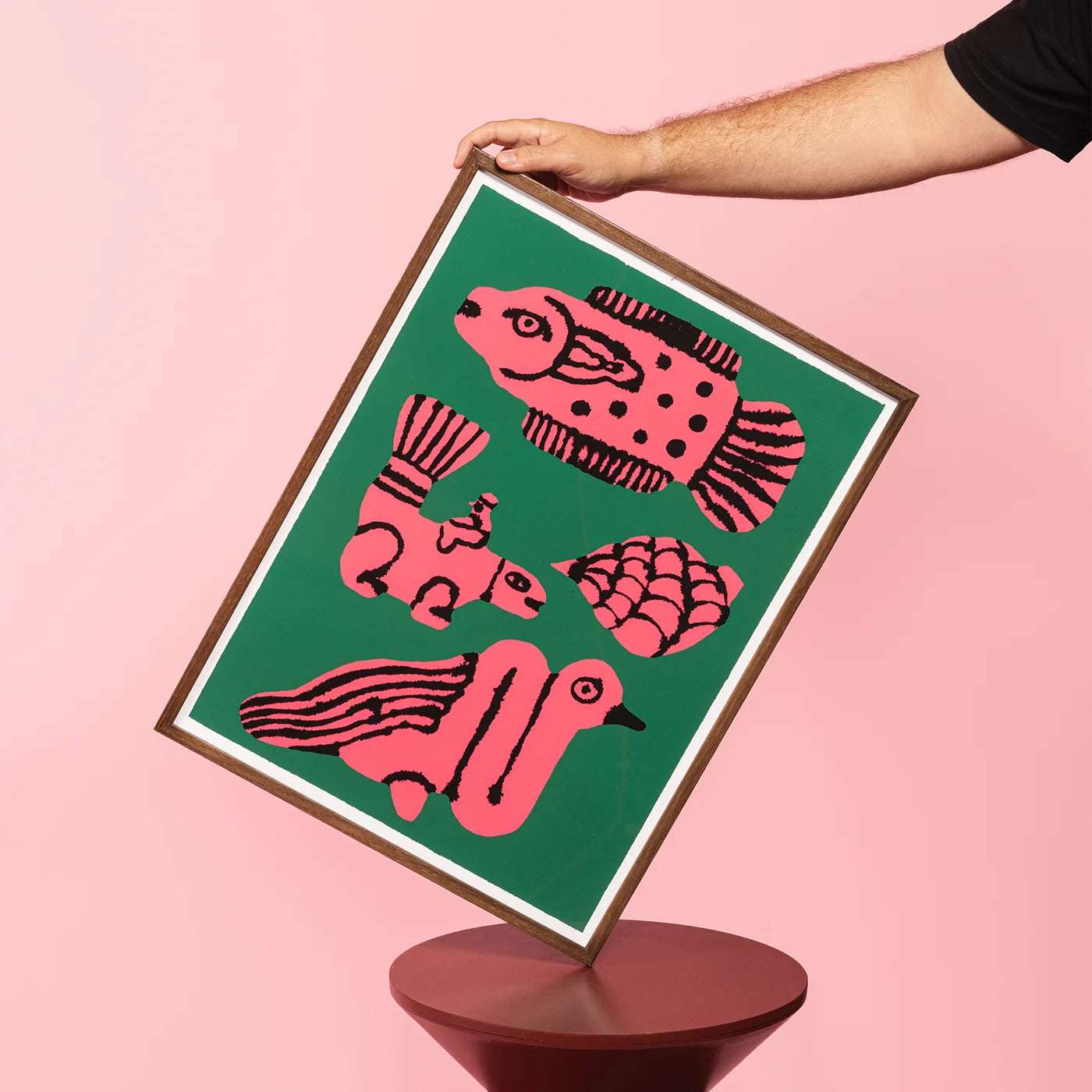A framed art print featuring stylized pink fish and duck illustrations on a green background, held by a person&