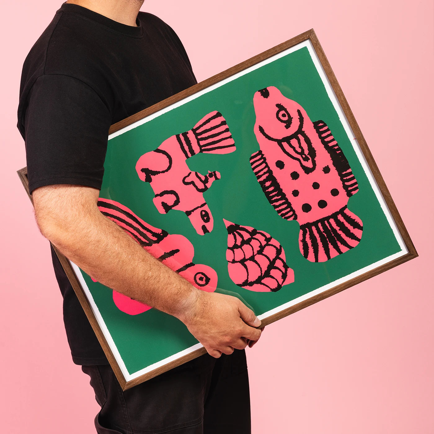 A person in a black t-shirt holds a framed art print featuring pink stylized fish and shell motifs on a green background, set against a solid pink backdrop.