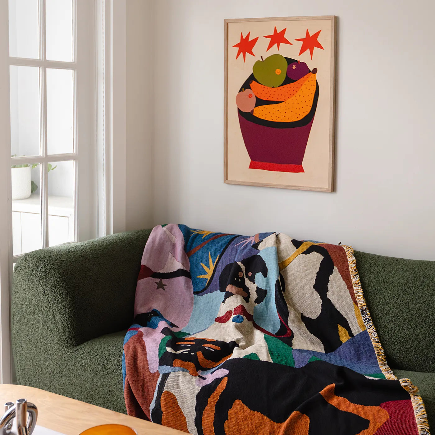 A colorful abstract-patterned throw blanket is draped over a dark green sofa in a living room setting, with a framed art print hanging on the wall above.