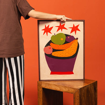 A framed art print featuring a colorful fruit bowl illustration with red stars, held by a person wearing a brown shirt and black-and-white striped pants against a solid orange background.