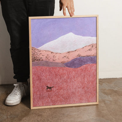 A framed art print depicting a painted landscape with a white mountain, pink and purple hills, and a small dark figure in a pink field, held by a person wearing black pants and white sneakers.