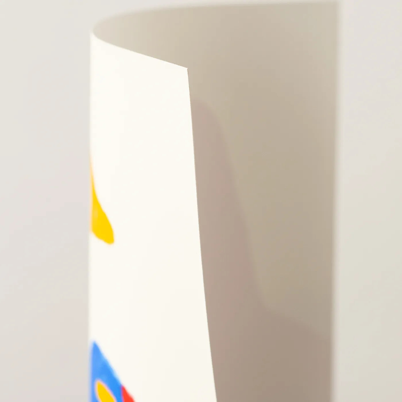 A close-up of a white art print with a curved corner, featuring a partial colorful graphic in yellow, blue, and red near the bottom, set against a plain light gray background.