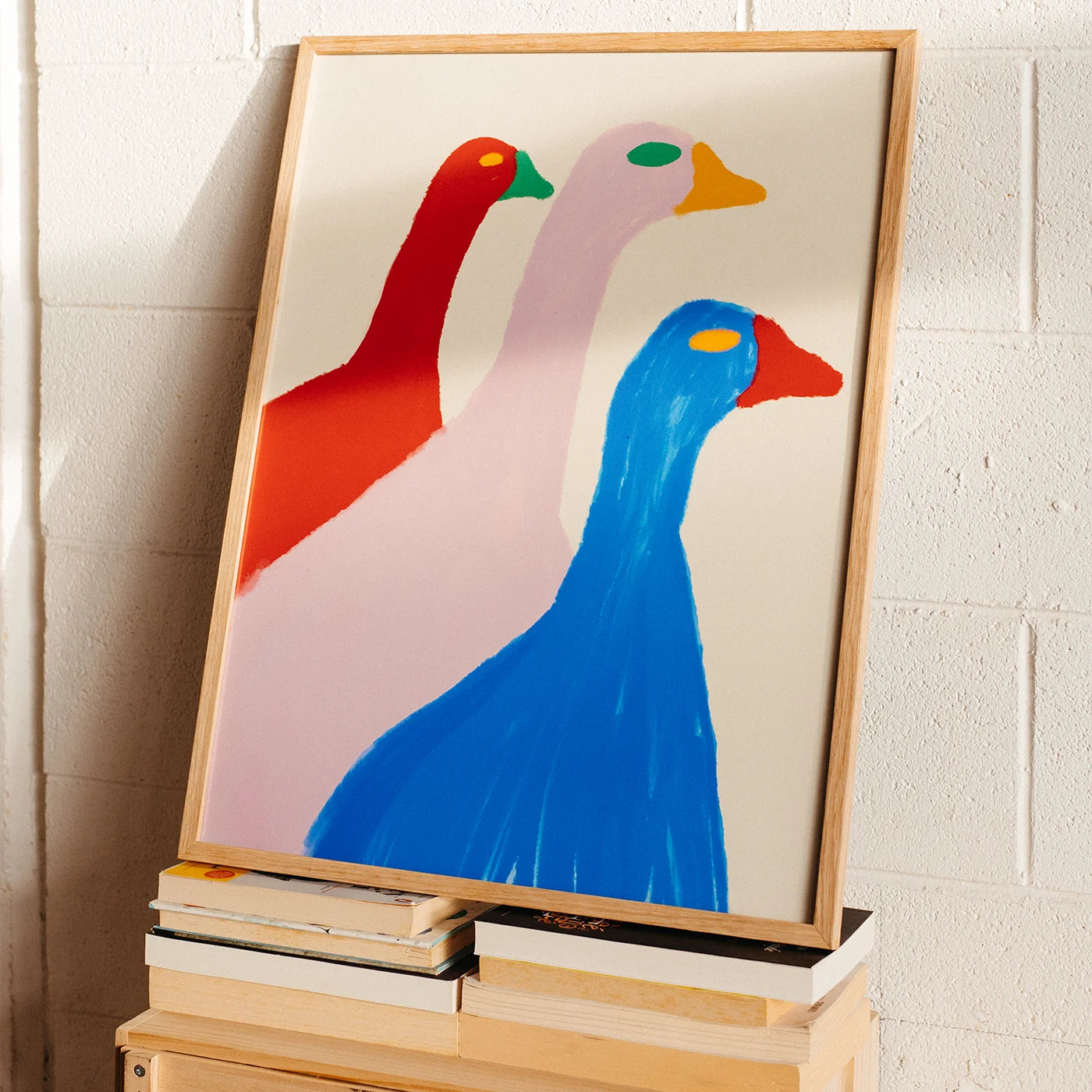 A framed abstract art print featuring three stylized birds in red, pale pink, and blue, leaning against a white brick wall on top of stacked books.