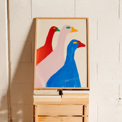 A framed abstract art print depicting three stylized geese in red, pale pink, and blue, displayed on a wooden easel against a white painted brick wall.