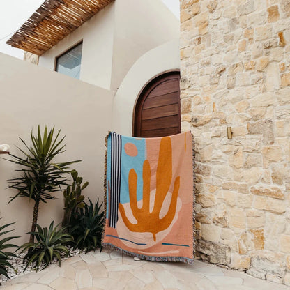 Woven throw blanket with cactus plant abstract design outside a sandstone home.