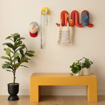 A styled entryway scene with a mustard yellow bench against a cream wall, a red Heller Swell Wall Catchall holding hats, a leash, and a tote bag, plus potted plants on the floor.