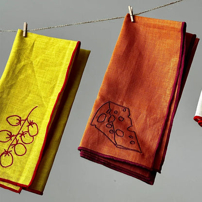 Two embroidered napkins, one yellow with red embroidery and one orange with a cheese design, hanging on a line against a gray background.