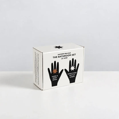 A white cardboard box for Maison &amp; Balzac The Bathroom Set Le Soin, featuring black text and hand graphics on the front, photographed on a clean white surface against a light gray background.