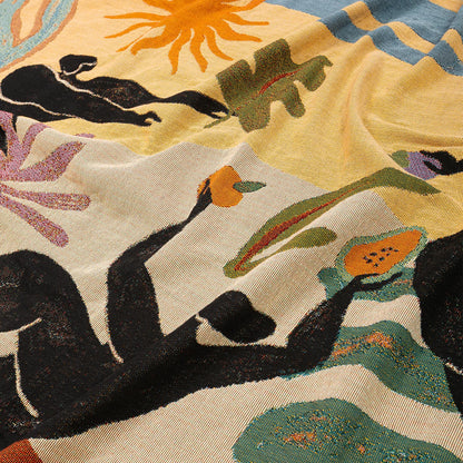 A close-up of a woven blanket featuring a colorful abstract and botanical pattern with black silhouetted figures, green leaves, orange sun and fruit, and purple floral elements on a cream and pale yellow background.