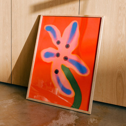 A framed abstract floral art print with a bright orange background, a pink flower shape outlined in yellow, blue petal-like strokes, and a green stem, leaning against a light wood cabinet on a concrete floor.