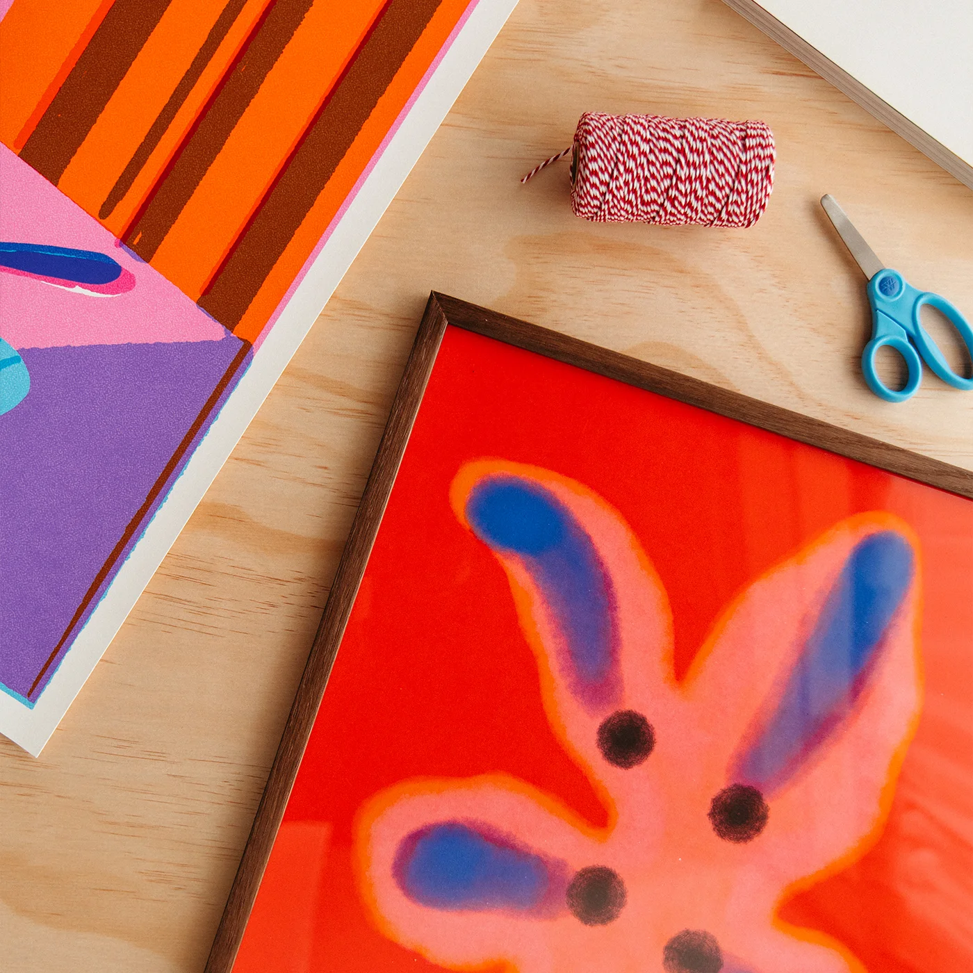 A framed abstract art print featuring a stylized flower in vibrant orange, pink, and blue on a light wood background, styled with craft supplies including a spool of red and white striped twine and blue-handled scissors.