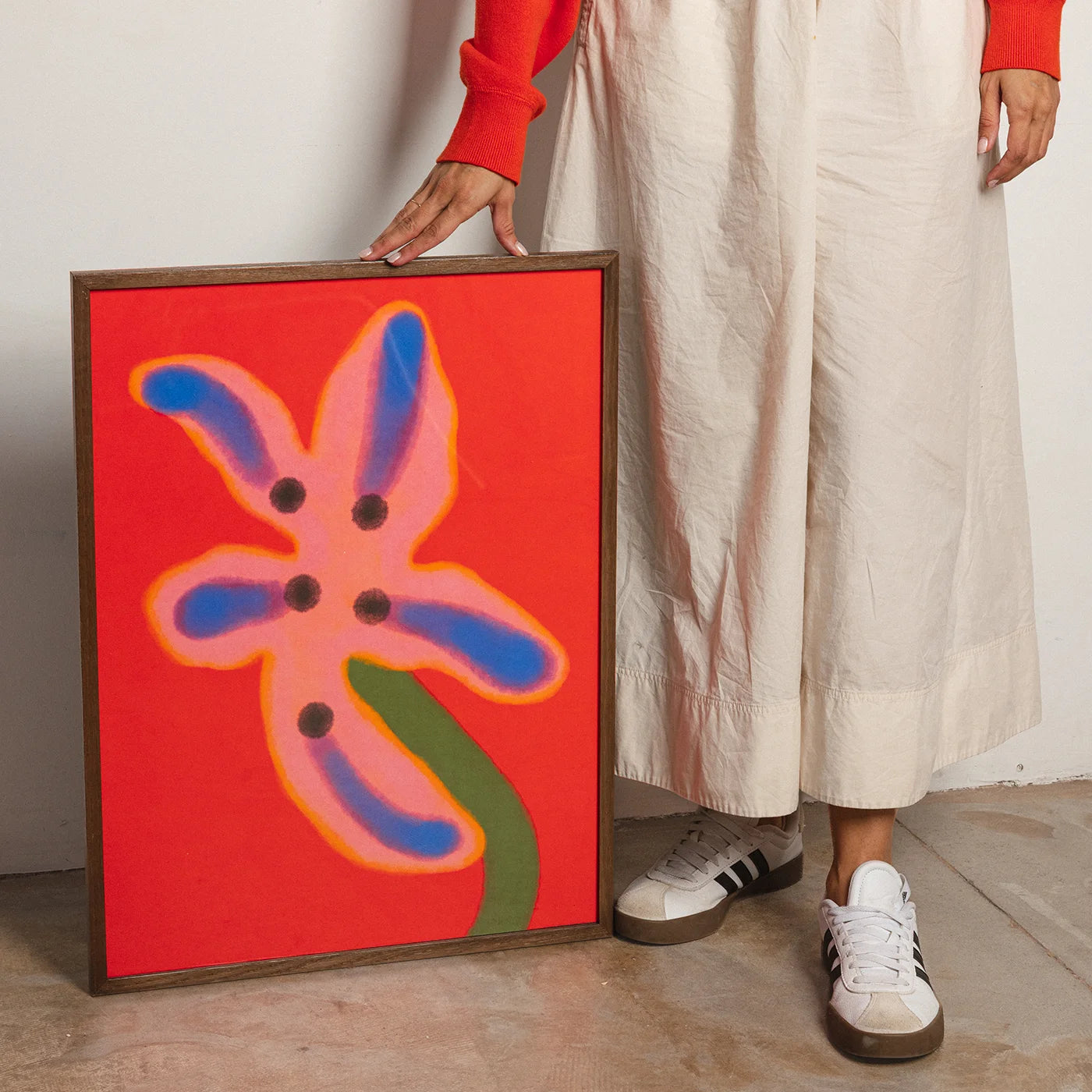 Framed abstract wall art with a bright red background, pink flower shape outlined in orange, blue petal-like shapes, and a green stem, leaning against a wall while a person in a cream skirt, red top, and white sneakers holds one side.