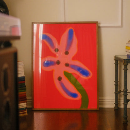 A framed abstract floral art print with a bright red background, pink flower shape, blue petal outlines, and a green stem, leaning against a cream wall in a room with wood floors and books.
