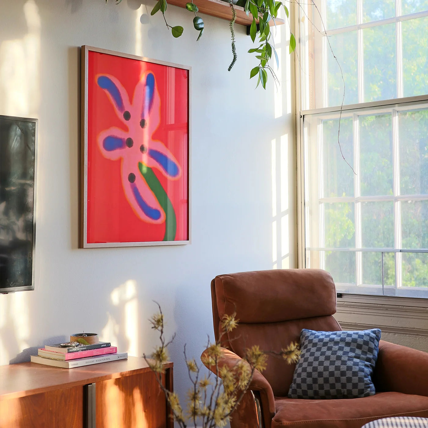 Wall art for living room depicting a framed abstract floral wall art print with a bright red background and pink, blue, and green flower-like shapes, displayed in a sunlit living room next to a brown armchair and window.