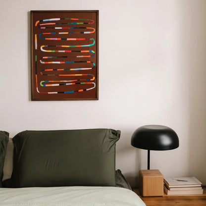 Wall art for bedroom featuring a framed abstract art print with colorful horizontal lines on a dark brown background, hanging on a light beige wall above a bed with green pillows and a black table lamp.