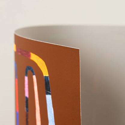 A close-up of an art print showing a brown background with abstract curved lines in yellow, pink, blue, and black, against a light neutral backdrop.