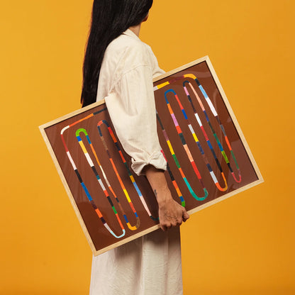 A person in a white shirt holds a large framed art print featuring colorful abstract ribbon-like lines on a brown background, set against a solid yellow backdrop.